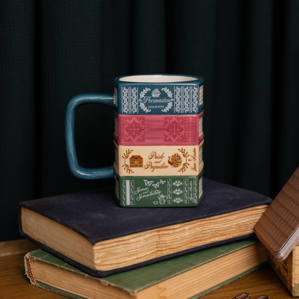 A ceramic mug designed to look like a stack of vintage hardcover books, featuring a teal handle and stacked book spines in blue, pink, beige, and green, displayed on a wooden table with old books and a dark curtain background.