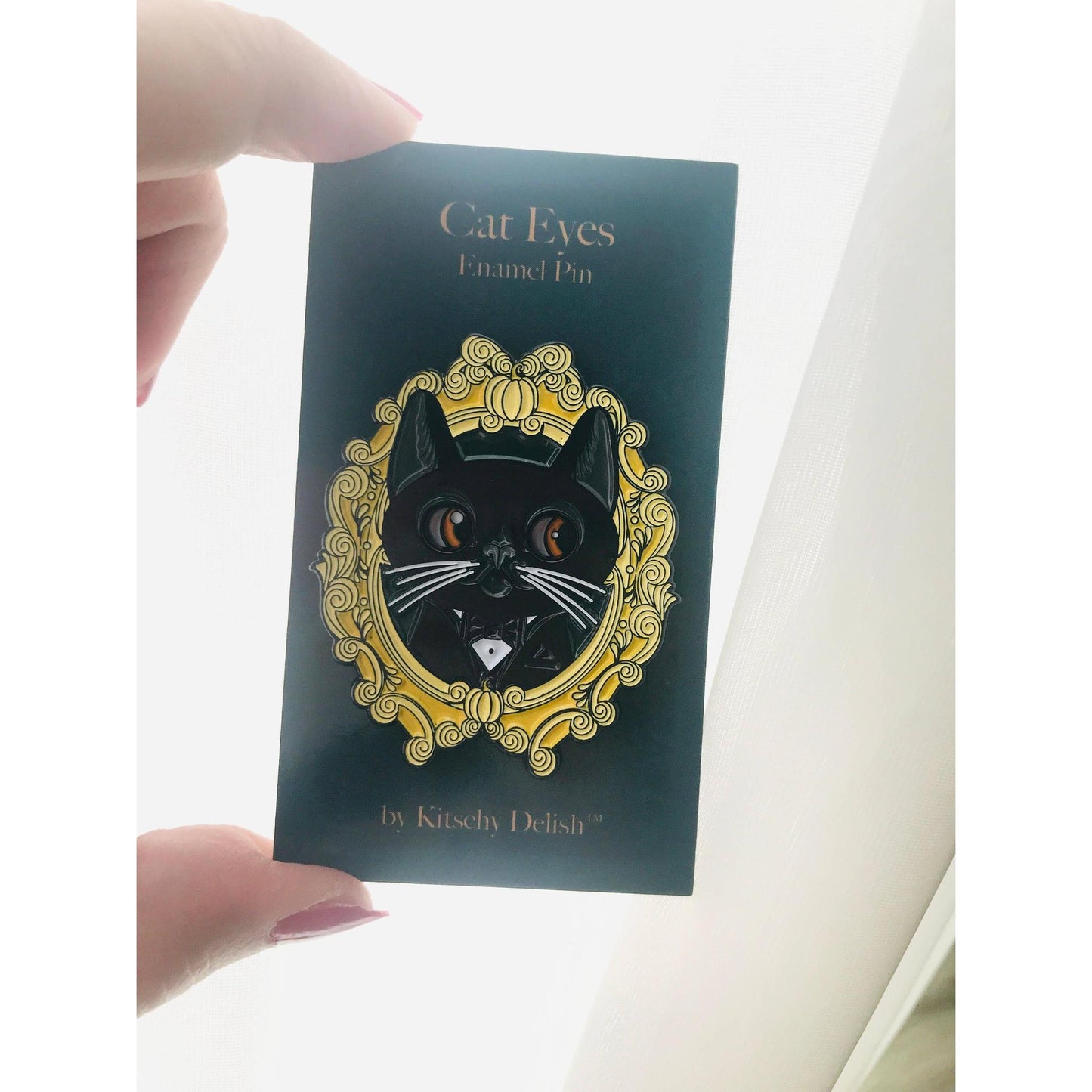 Enamel pin with cat design on a dark green card held by a hand.