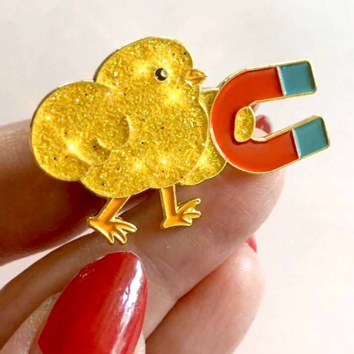 Gold chick-shaped magnet pin held by a hand with red nail polish on a light background