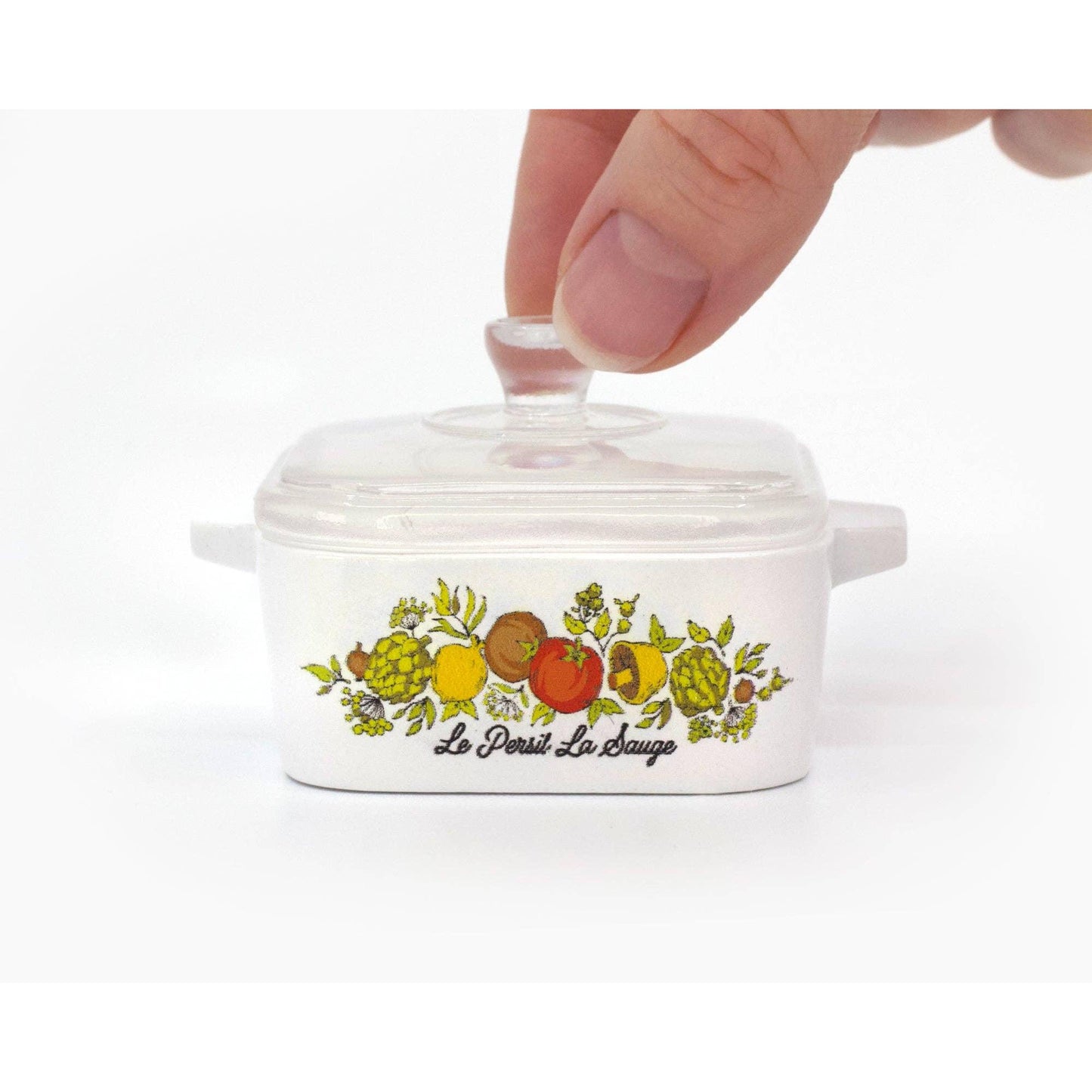 Small white ceramic container with fruit design and 'Le Persit La Sauge' text, being held by a hand.