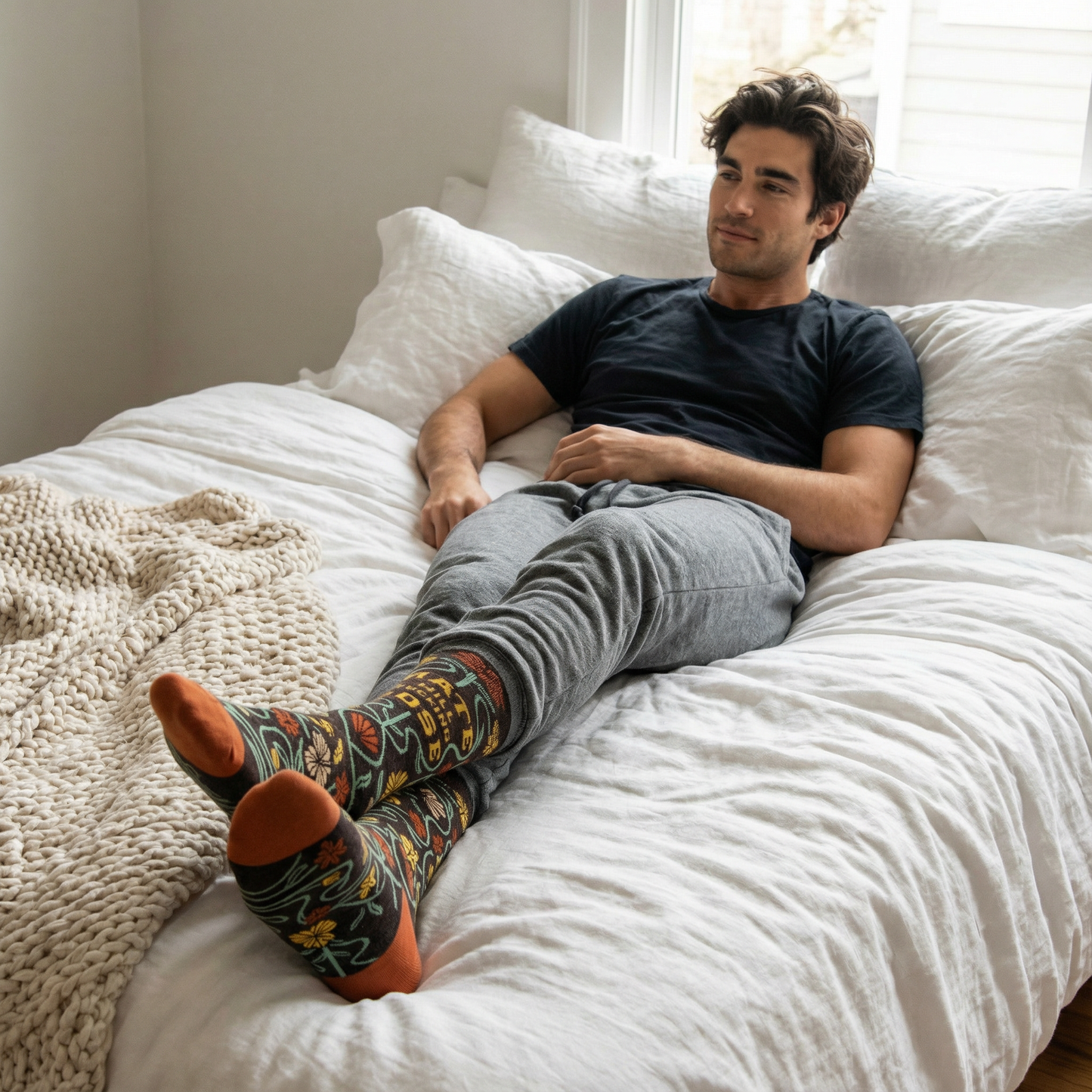 Man sitting on a bed wearing floral patterned socks and a black t-shirt.
