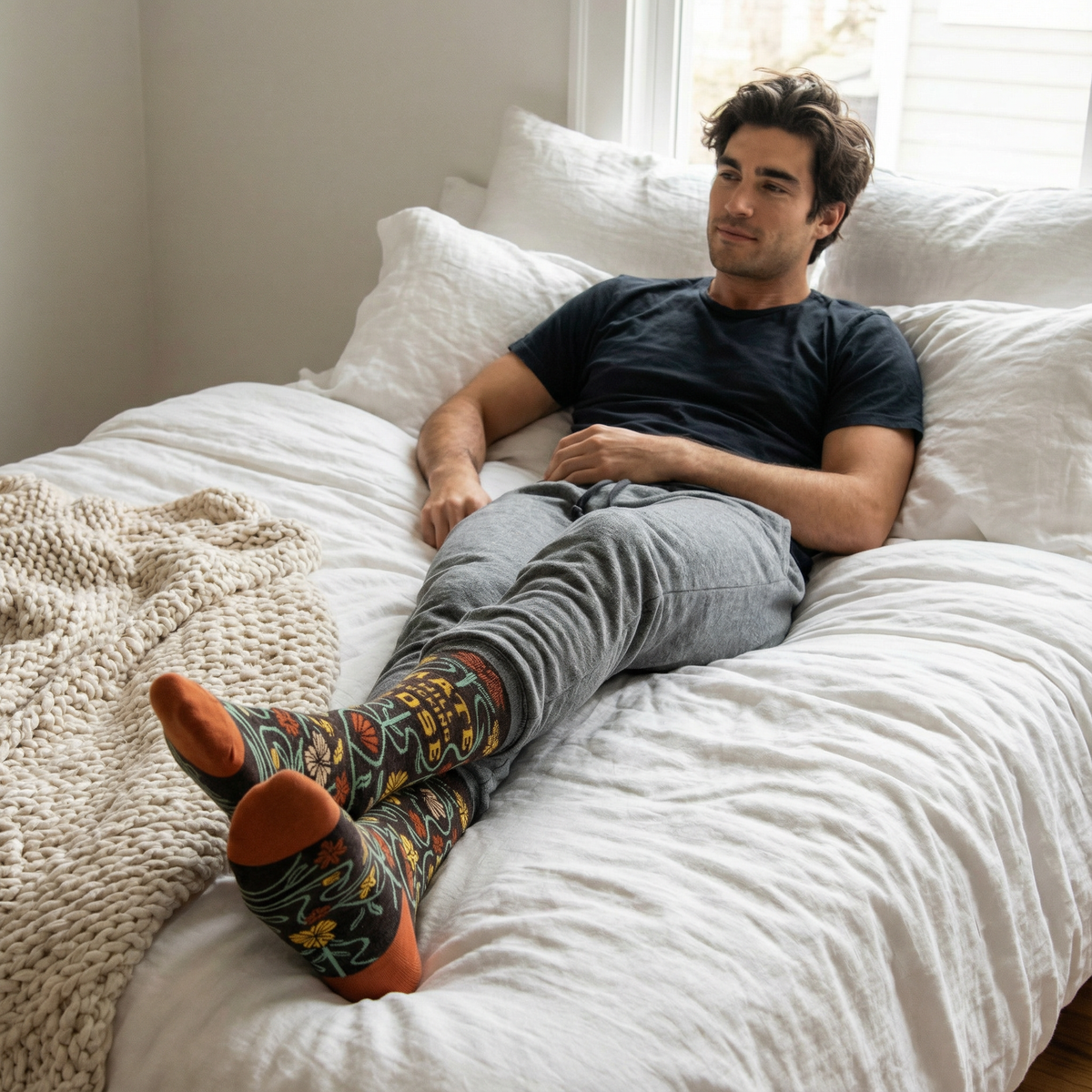 Man sitting on a bed wearing floral patterned socks and a black t-shirt.
