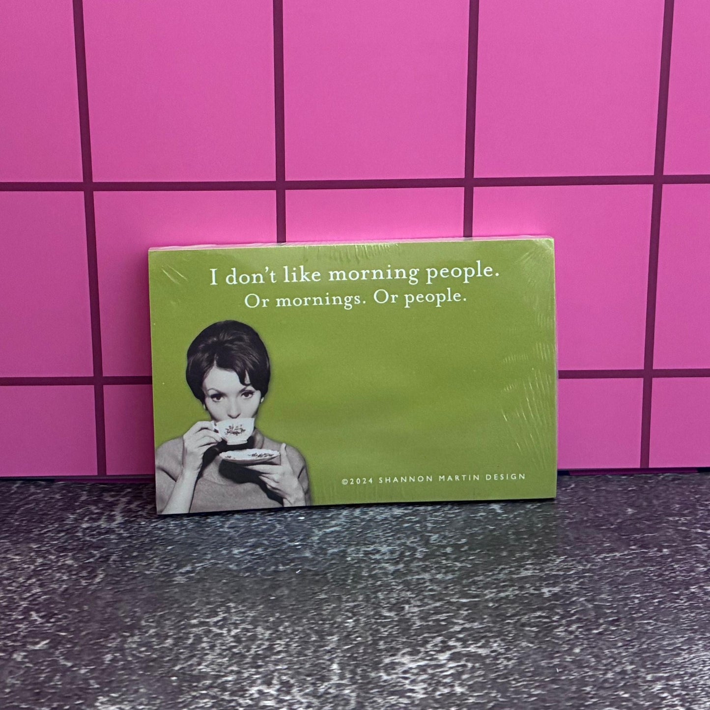 Green sticky note pad with retro lady drinking cofee and text on a pink tiled wall.