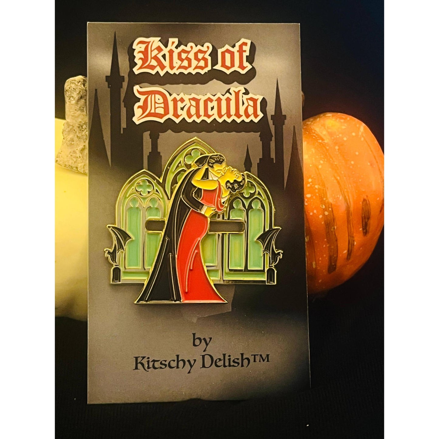 Enamel pin of a couple in front of a castle with 'Kiss of Dracula' text on a card, surrounded by pumpkins.