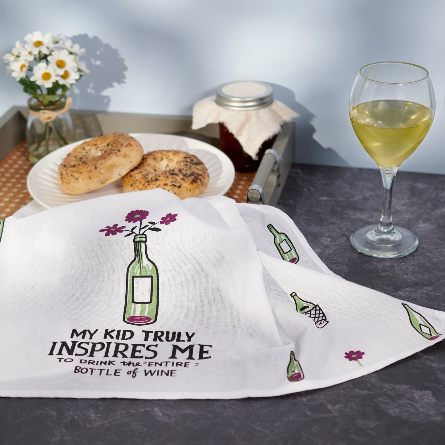 White towel with wine bottle design and text, placed on a table with bagels and a glass of wine.