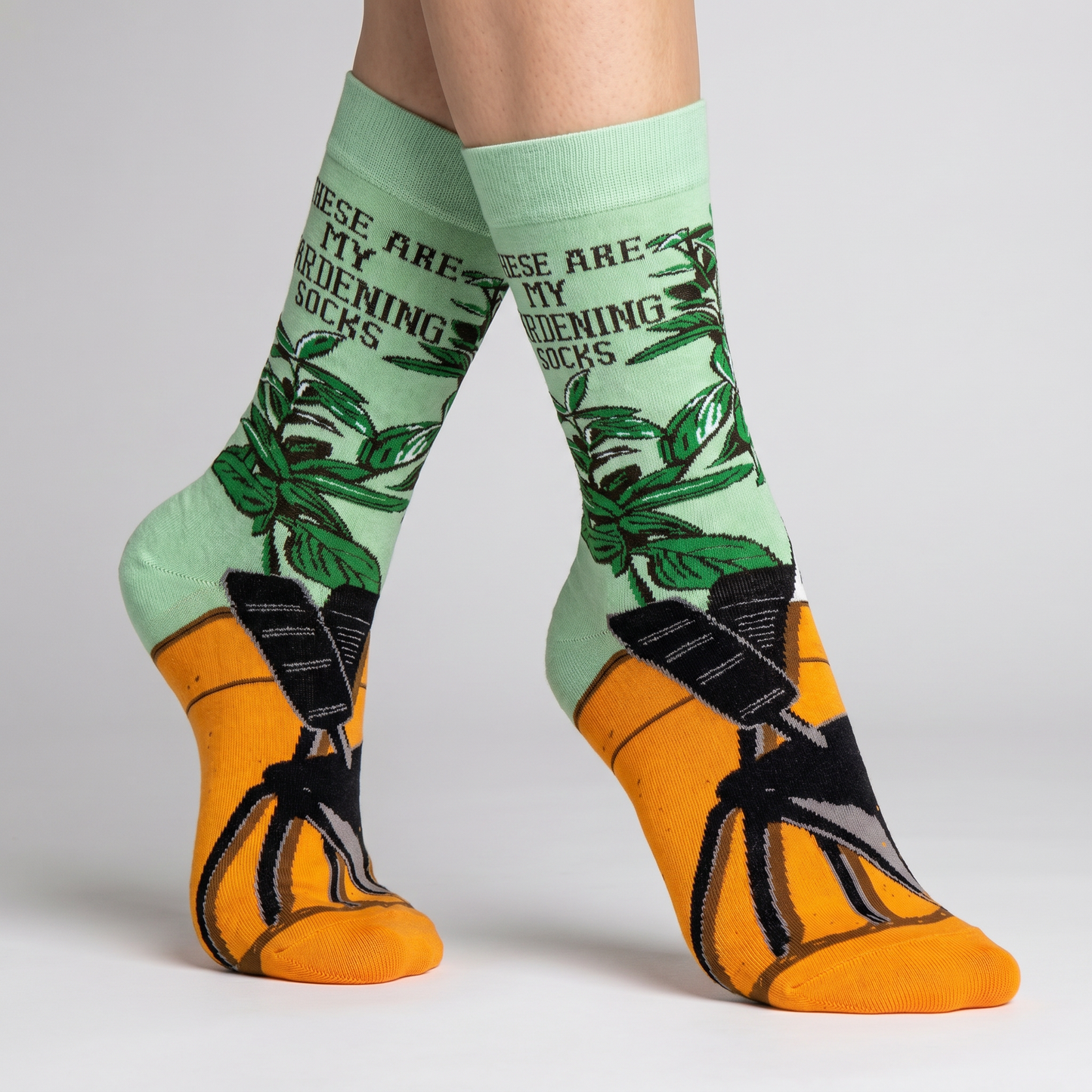 Green gardening-themed socks with text and black and orange socks worn by a person on a light gray background.
