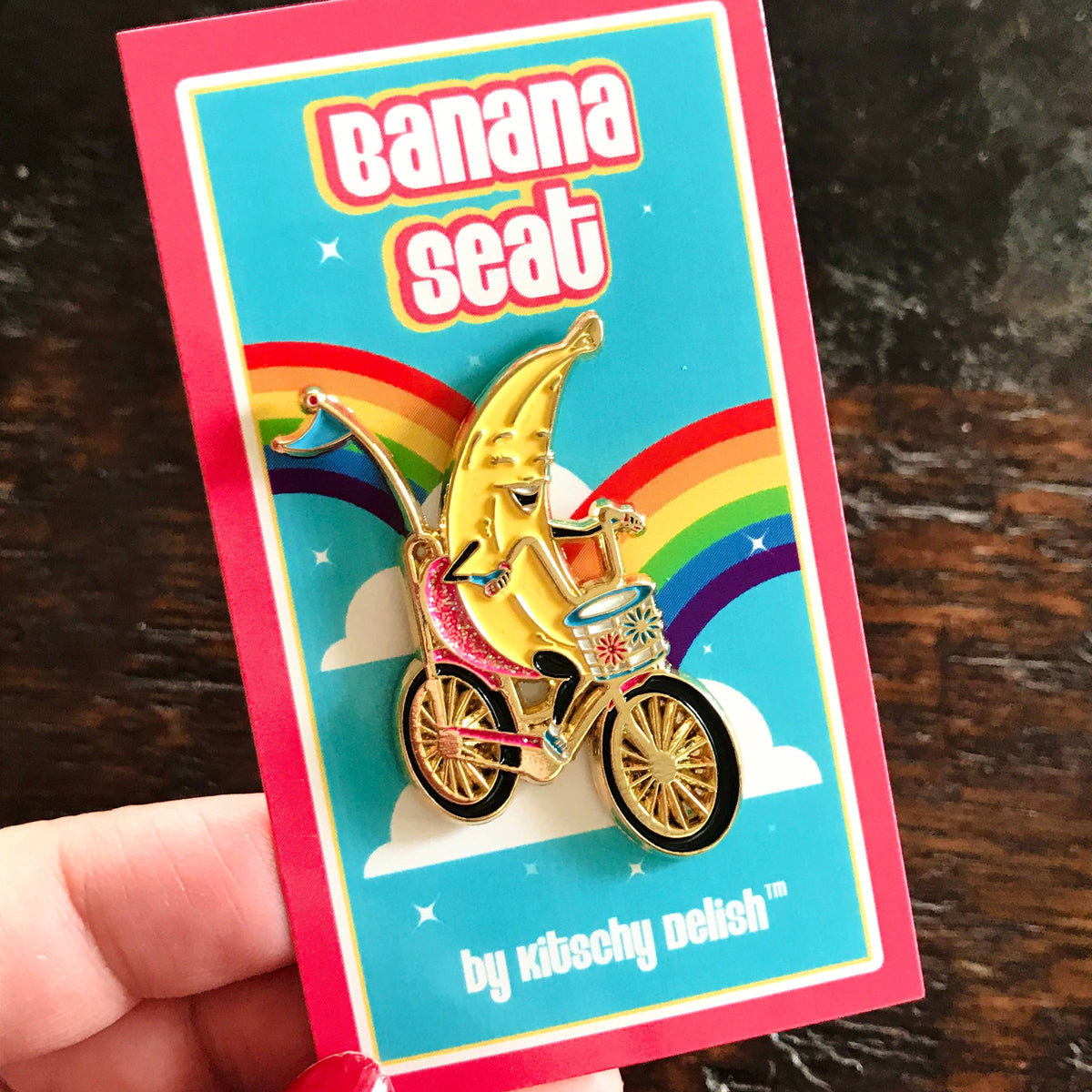 A hand holds a colorful enamel pin on its backing card. The pin features a smiling banana riding a bicycle with a basket, set against a rainbow and clouds on a turquoise background.