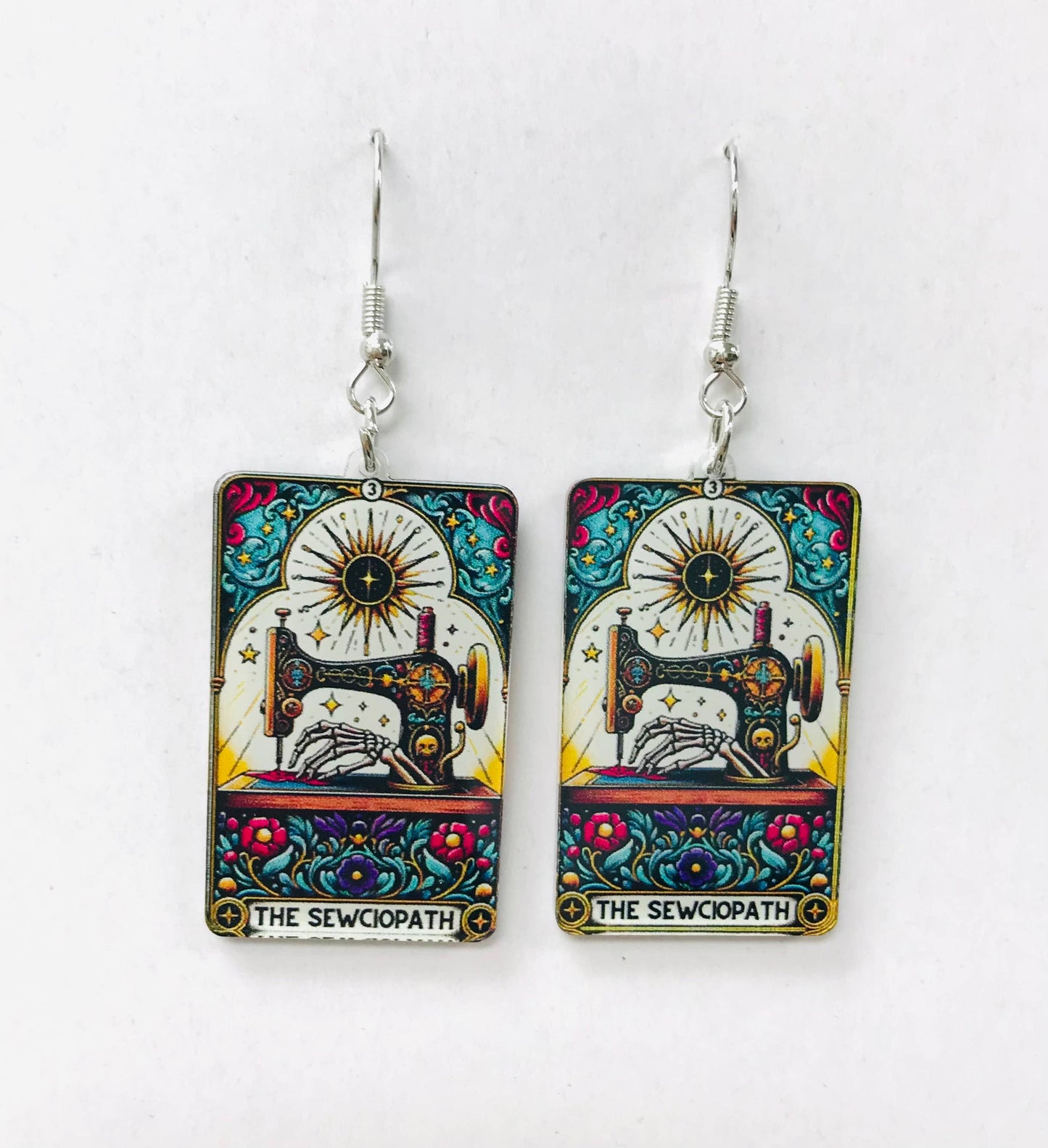 Colorful earrings with a sewing machine design and The Sewciopath text