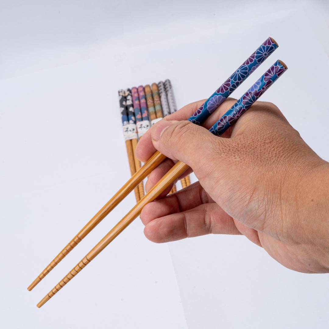 A hand holding a pair of bamboo chopsticks with blue and pink patterned handles, with several other colorful chopsticks bundled in the background against a plain white backdrop.