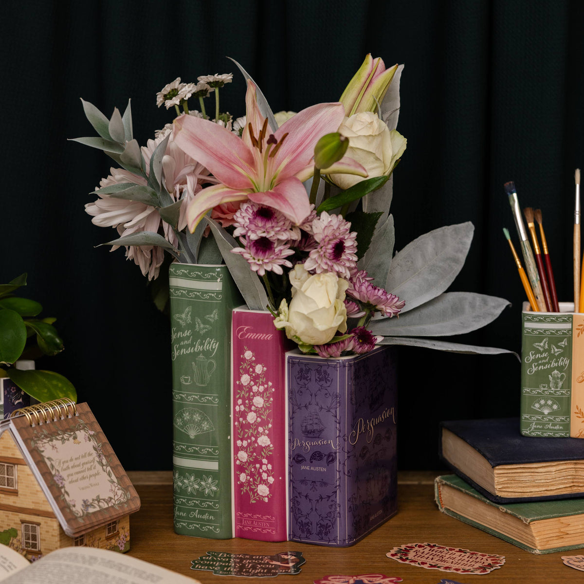Three vintage-style book spines used as vases containing a pink lily, white roses, and purple flowers, styled on a wooden desk with books, paintbrushes, and stickers against a black curtain background.