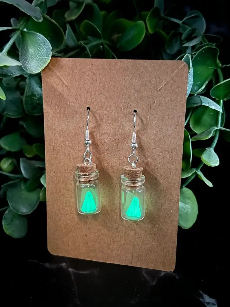 Glowing green jar earrings on a brown card with green leaves in the background