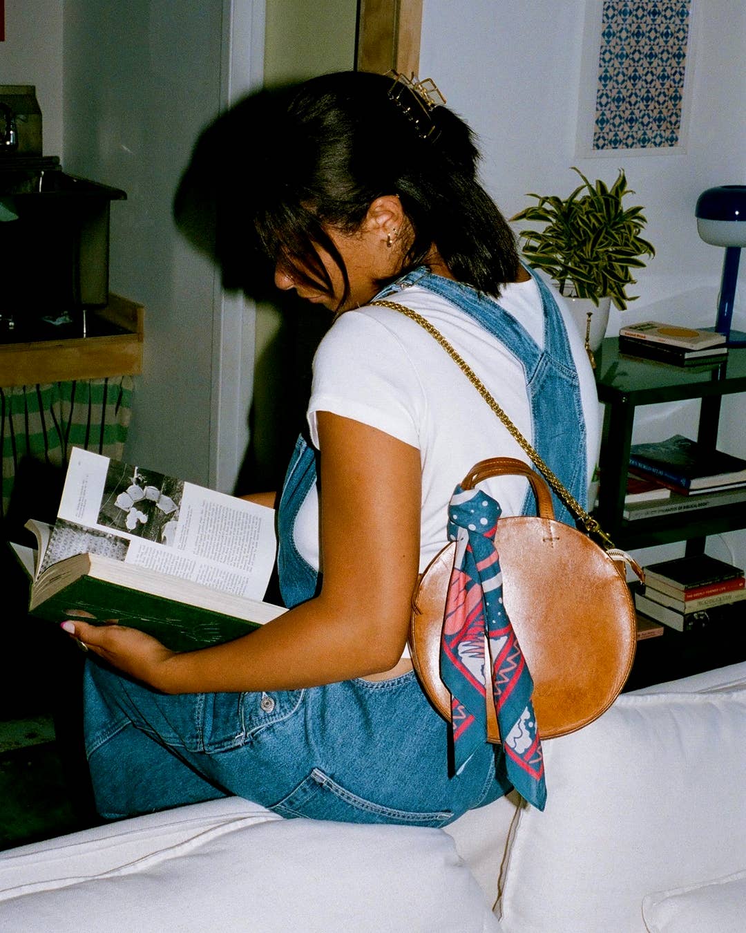 Woman sitting on a couch reading a book with a brown bag over her shoulder with bandana as bag accessory