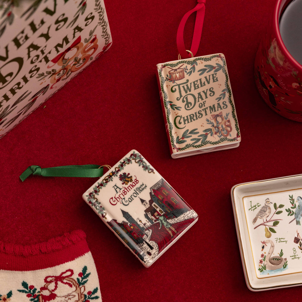 A styled flat-lay of Christmas-themed ceramic ornaments on a deep red fabric background, alongside a gift box, a red mug, and a stocking.