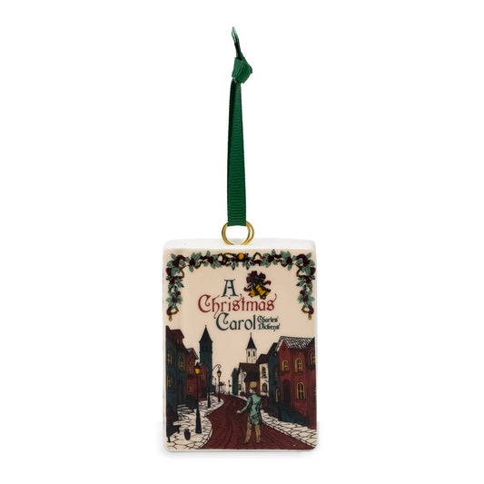 A square Christmas ornament featuring an illustrated winter street scene from 'A Christmas Carol' with a green hanging ribbon and gold loop.