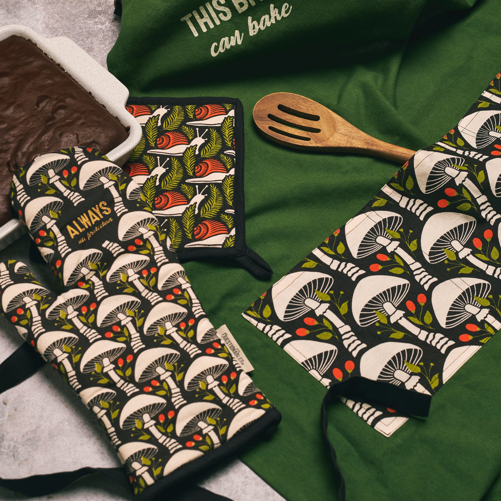 Mushroom-patterned oven mitts and pot holder on a green surface with a wooden spoon.