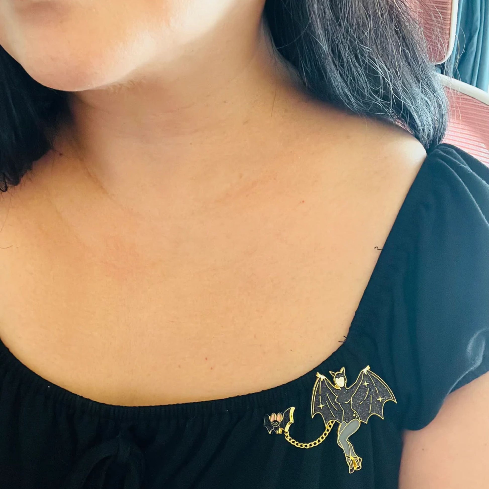 Person wearing a black top with a decorative bat enamel pin on the shoulder.