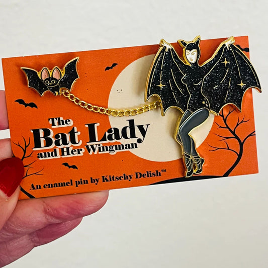 Enamel pin of a bat lady and her wingman on an orange card with text