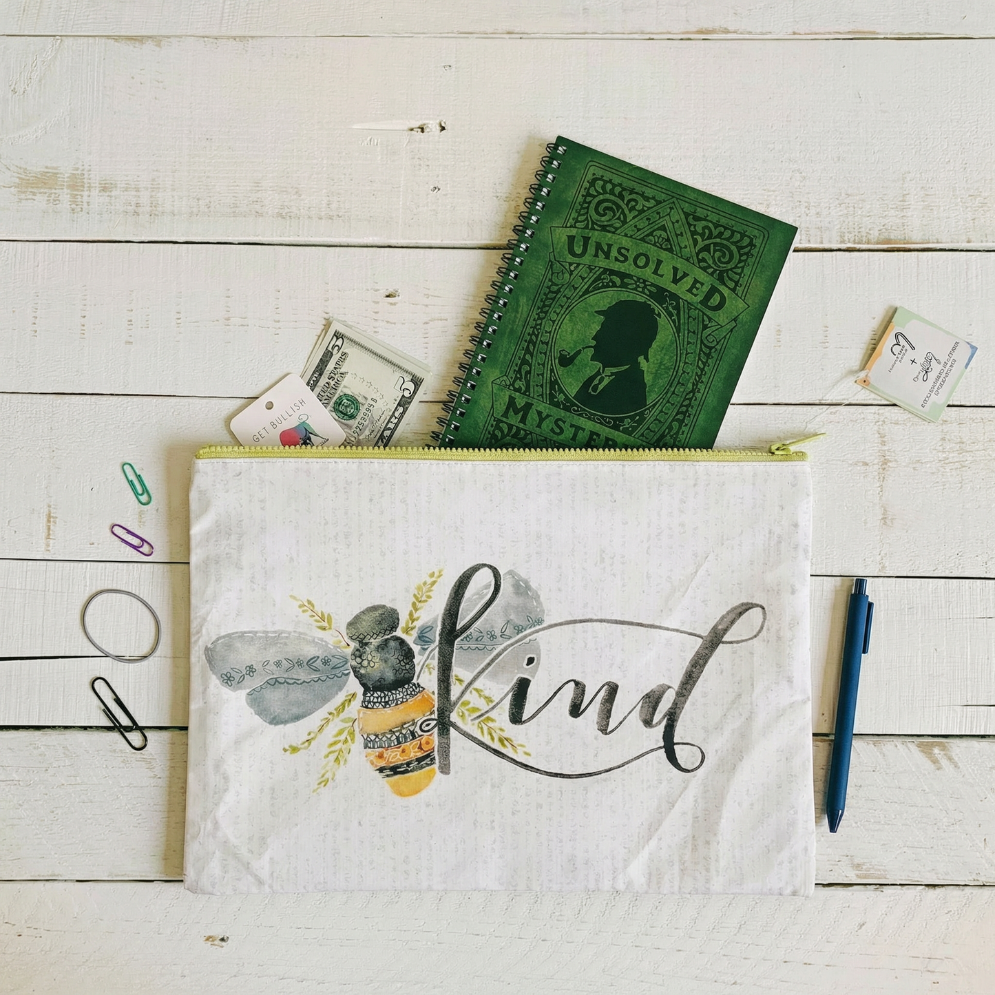 White pouch with bee design and 'Kind' text, green notebook, and money on a wooden surface.