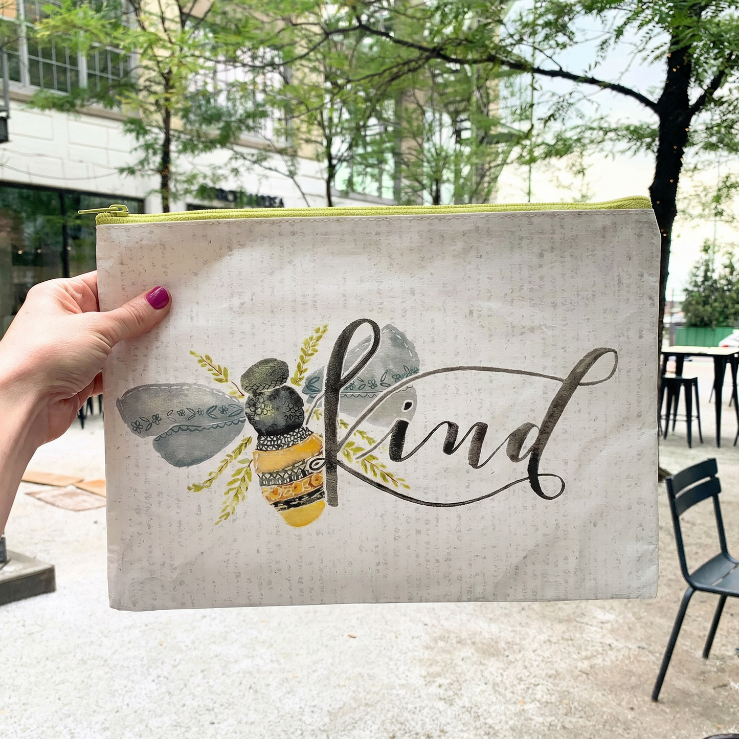 Hand holding a pouch with a bee design and 'Kind' text outdoors.