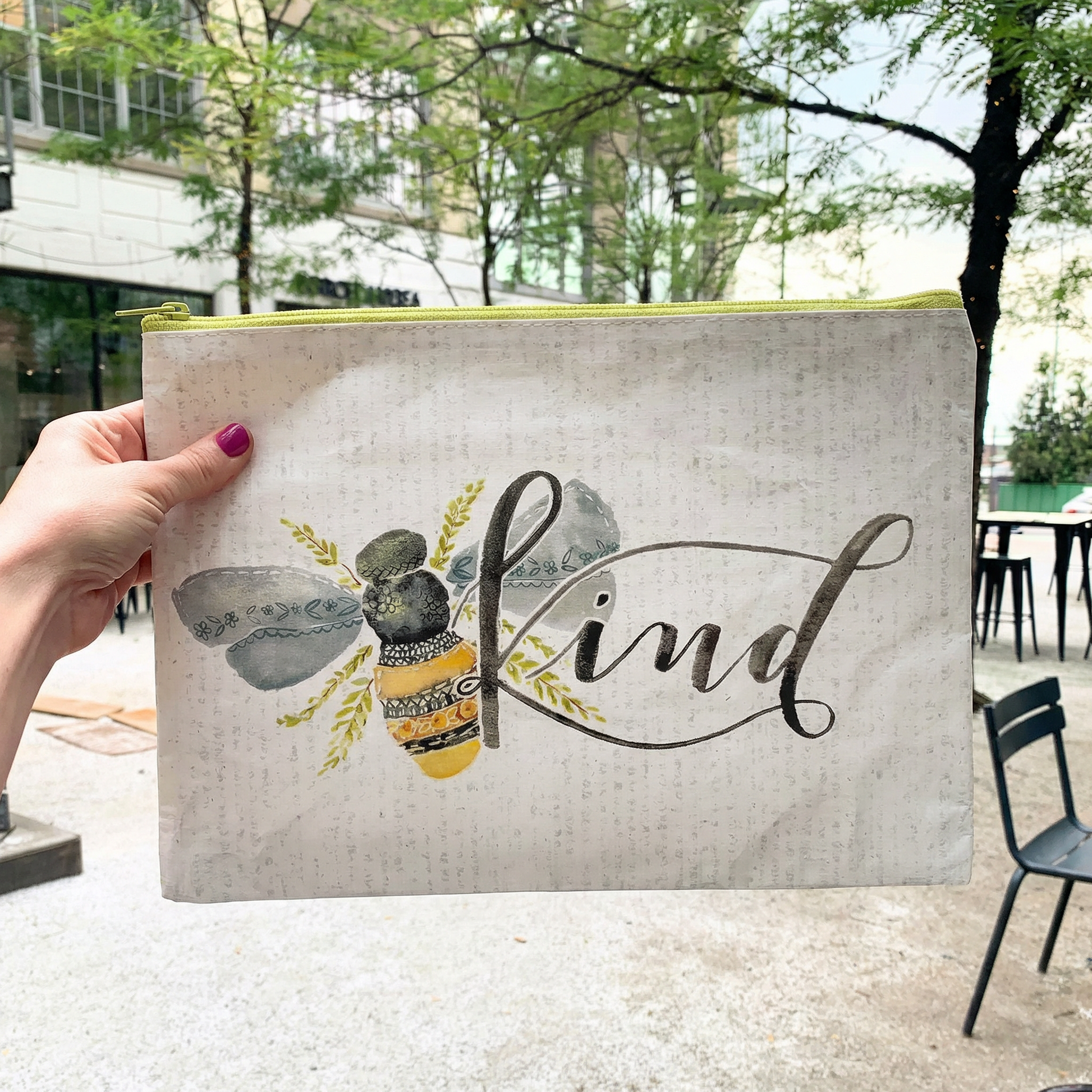 Hand holding a pouch with a bee design and 'Kind' text outdoors.