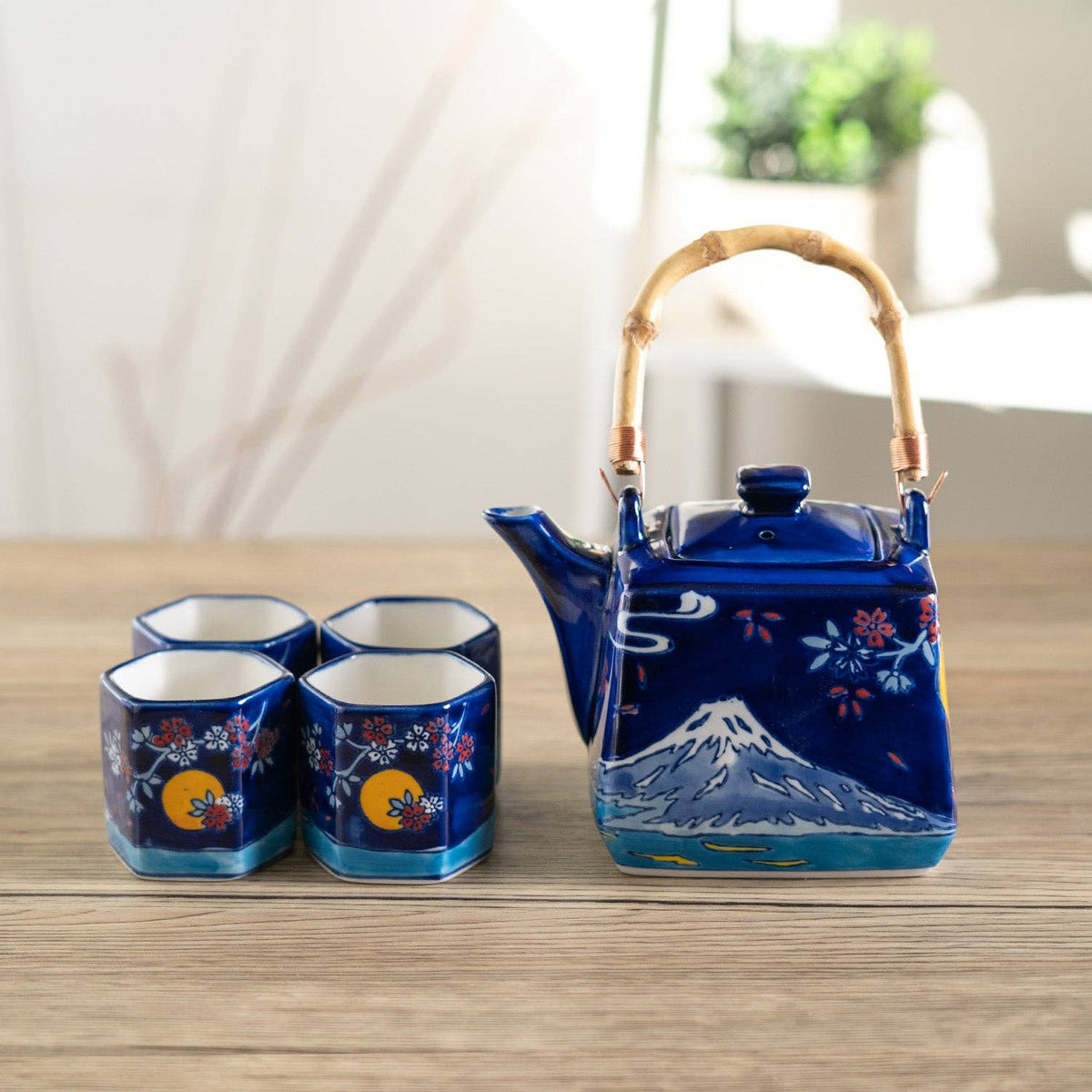 A ceramic tea set on a light wood tabletop, featuring a deep blue teapot with a bamboo handle and four matching small cups, all decorated with floral and mountain motifs.