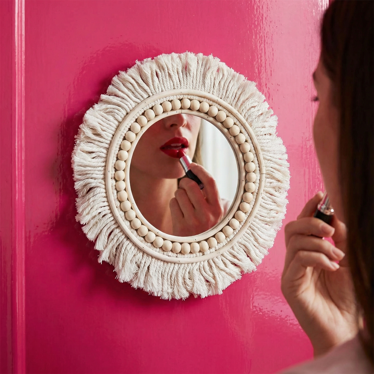 Person applying lipstick in front of a boho-inspired mirror with a pink background