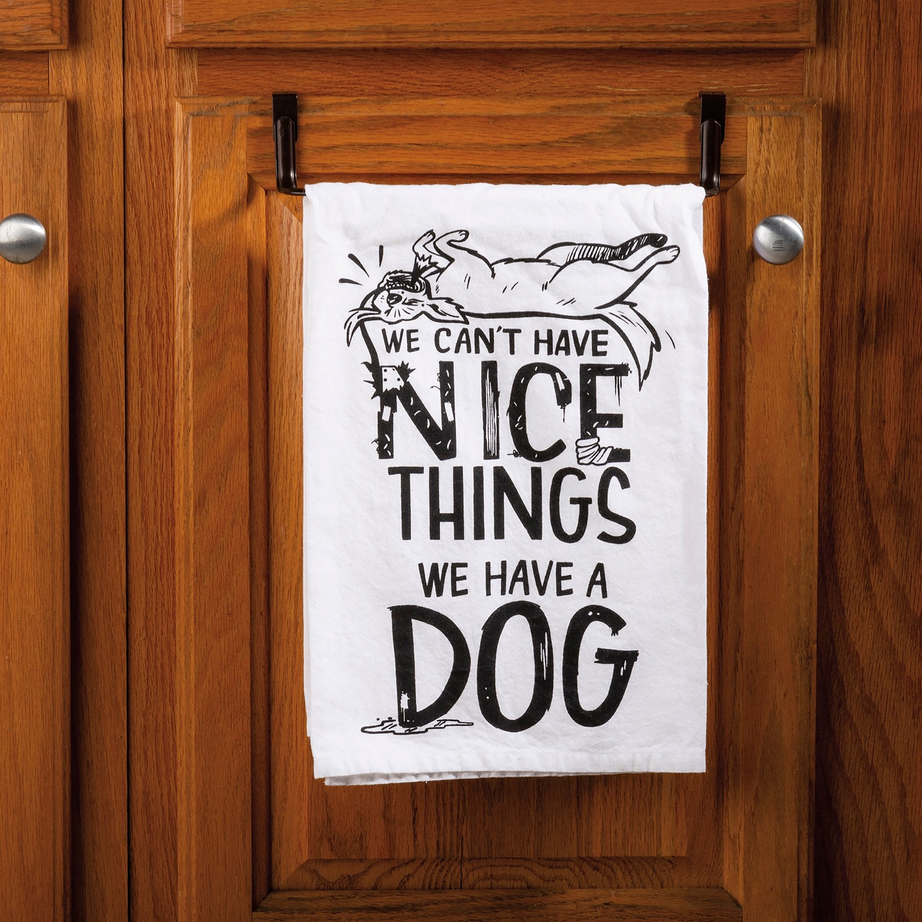 Tea towel with humorous text and dog illustration hanging on a wooden cabinet.