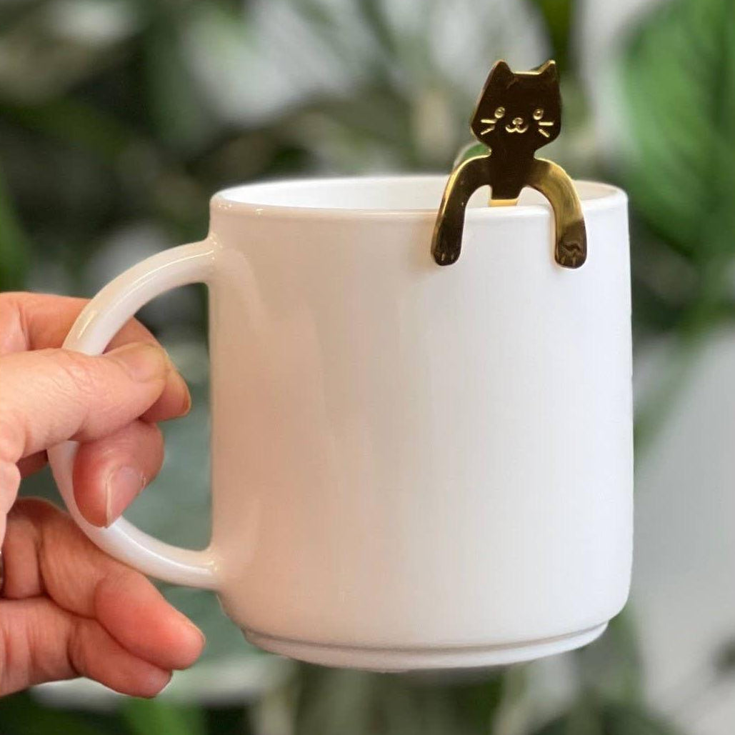 White mug with a gold cat-shaped handle held by a hand against a blurred green background