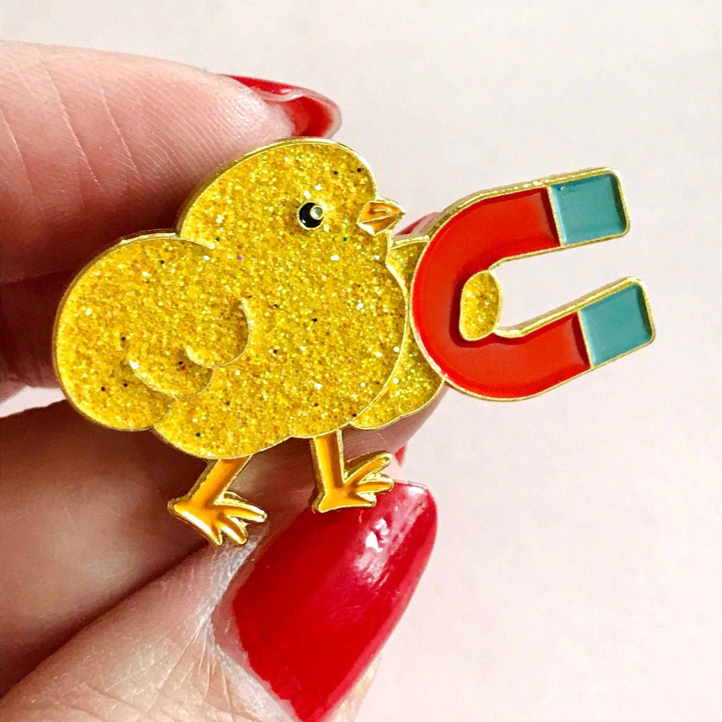 Gold chick-shaped magnet held between fingers with red nail polish on a light background