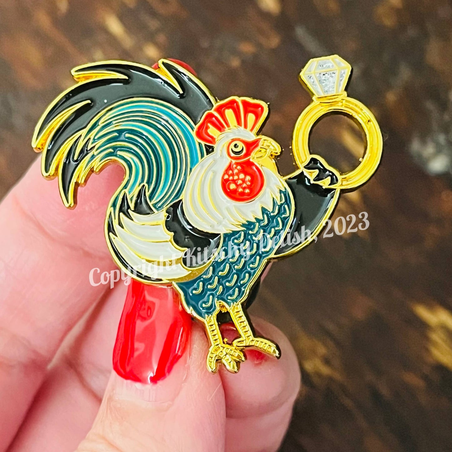 Colorful rooster-shaped enamel pin held by a hand with red nail polish.