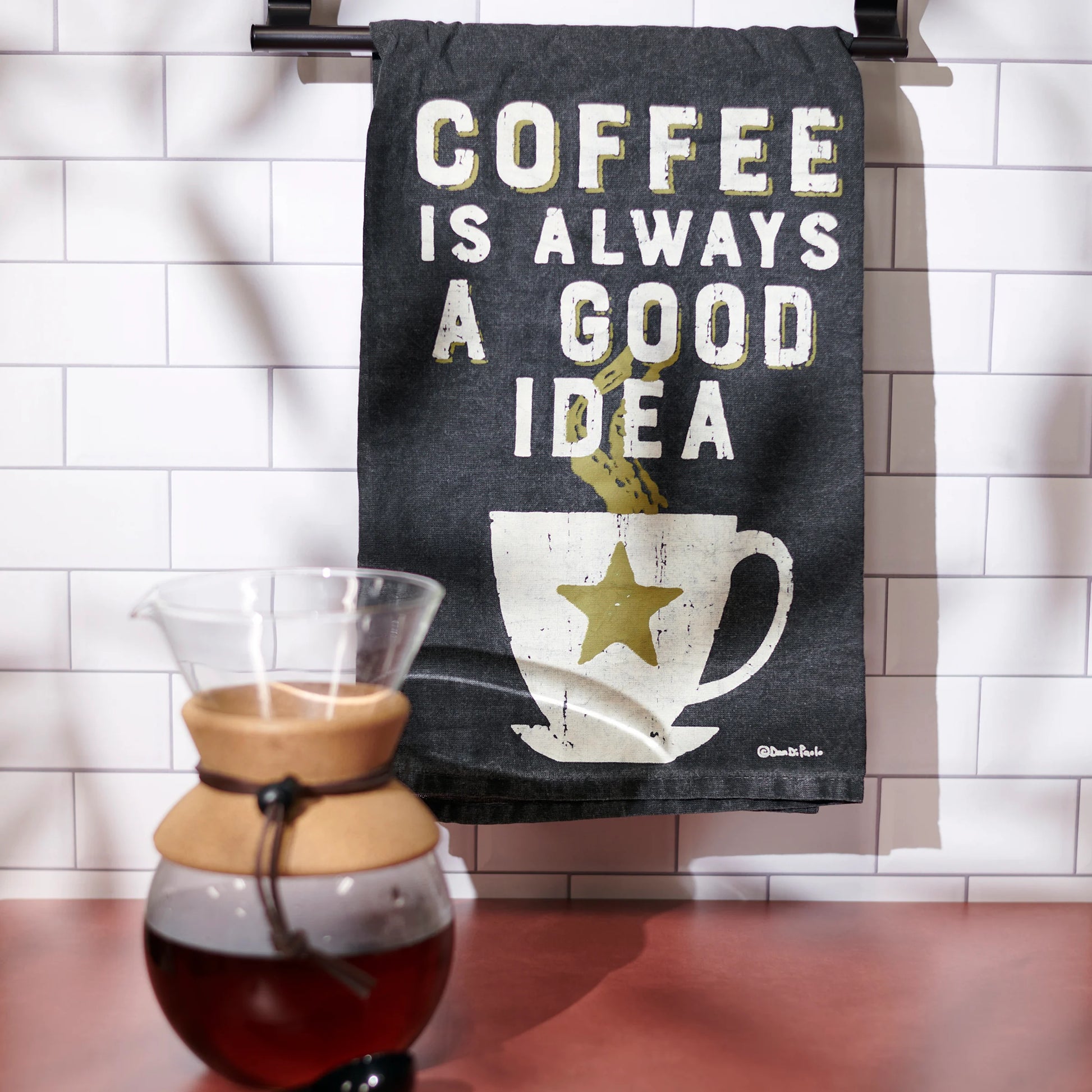 Towel with 'Coffee is always a good idea' text and coffee-themed design hanging on a rack, with a coffee carafe in the foreground.