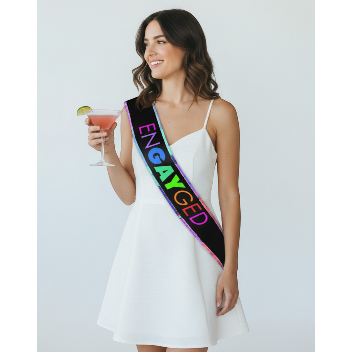 Woman in a white dress with a colorful 'ENGAYGED' sash, holding a cocktail.
