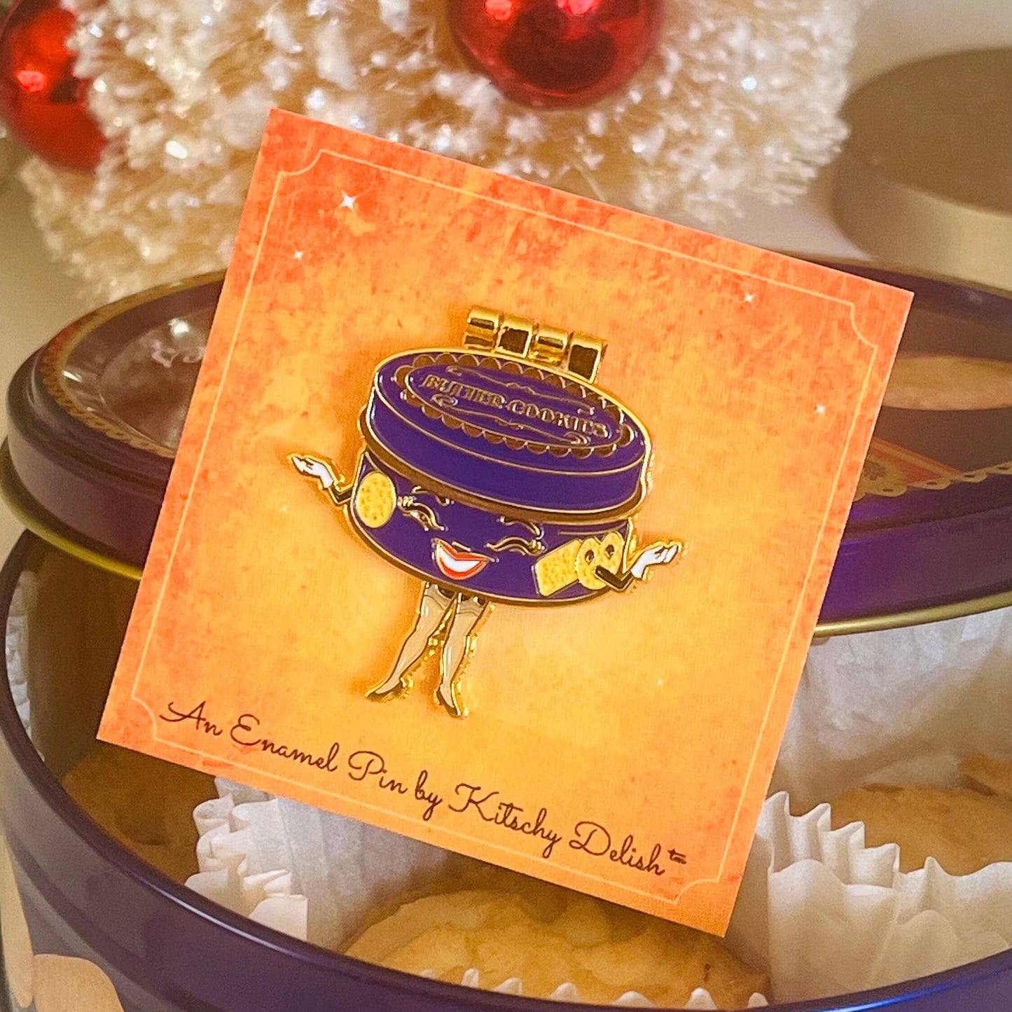 Enamel pin by Kitschy Delish on a decorative card with a Christmas decor in the background