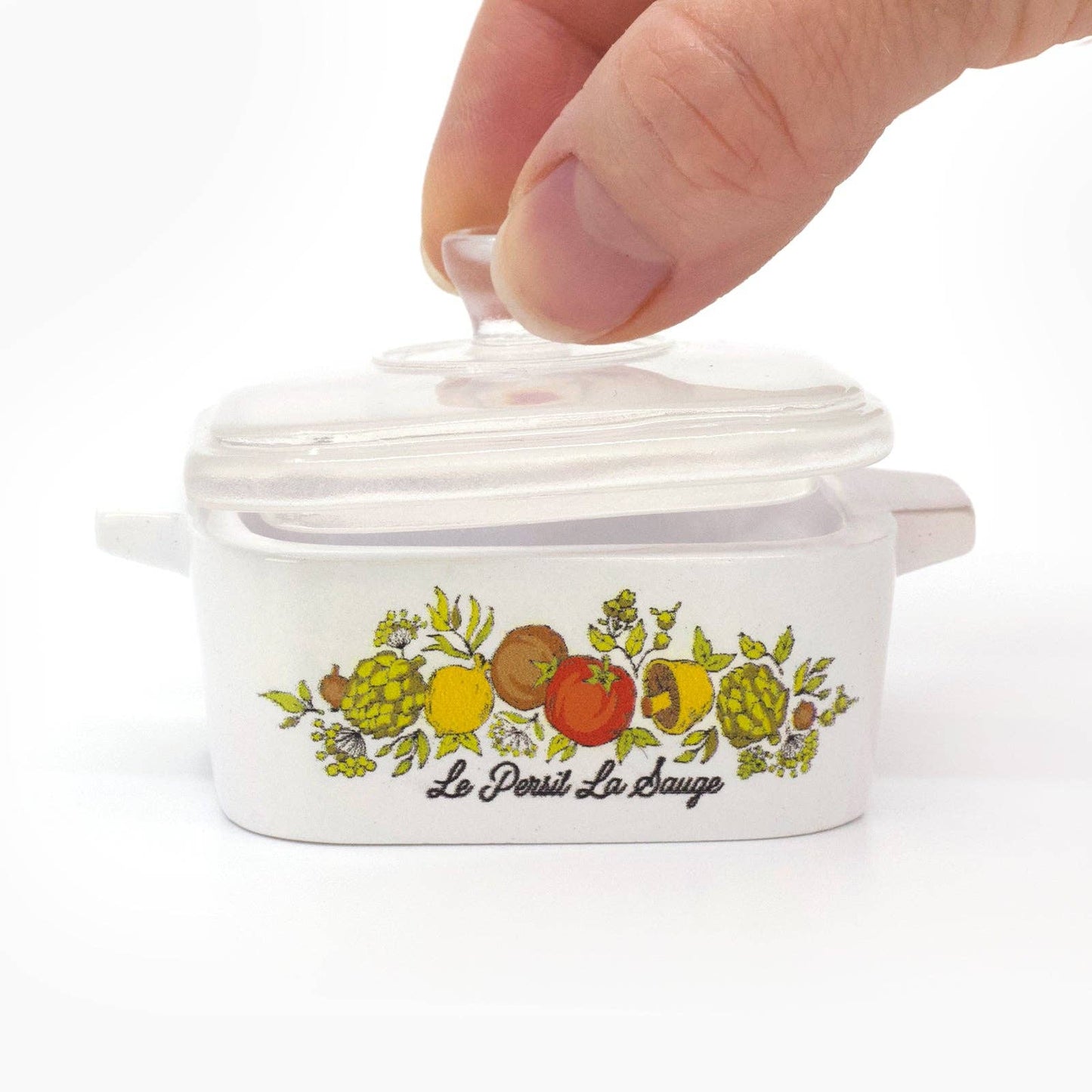 Hand opening a Corning Ware–Inspired Casserole with fruit design and text on a white background