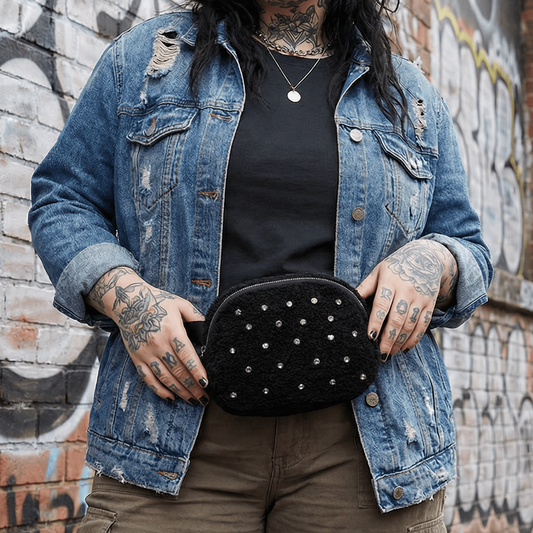 Person wearing a denim jacket and holding a black studded bag against a graffiti-covered wall.