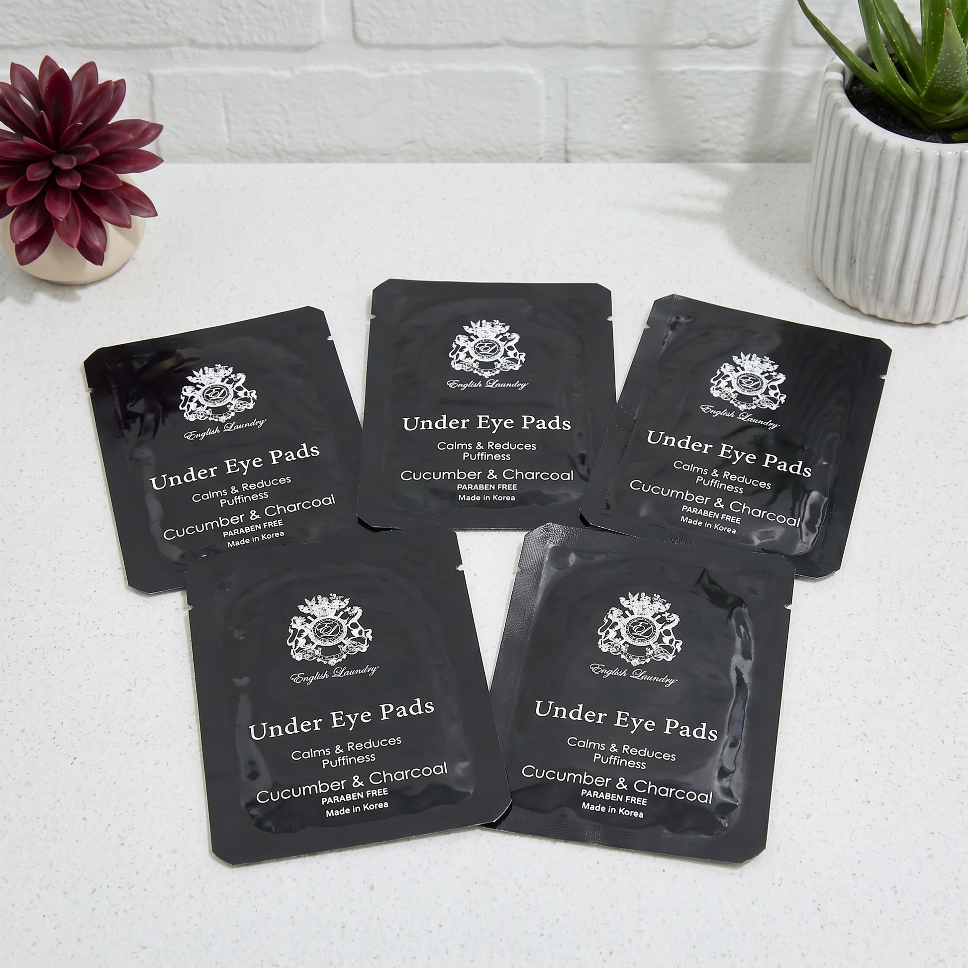 Five packages of Under Eye Pads with Cucumber & Charcoal on a white surface with decorative plants.