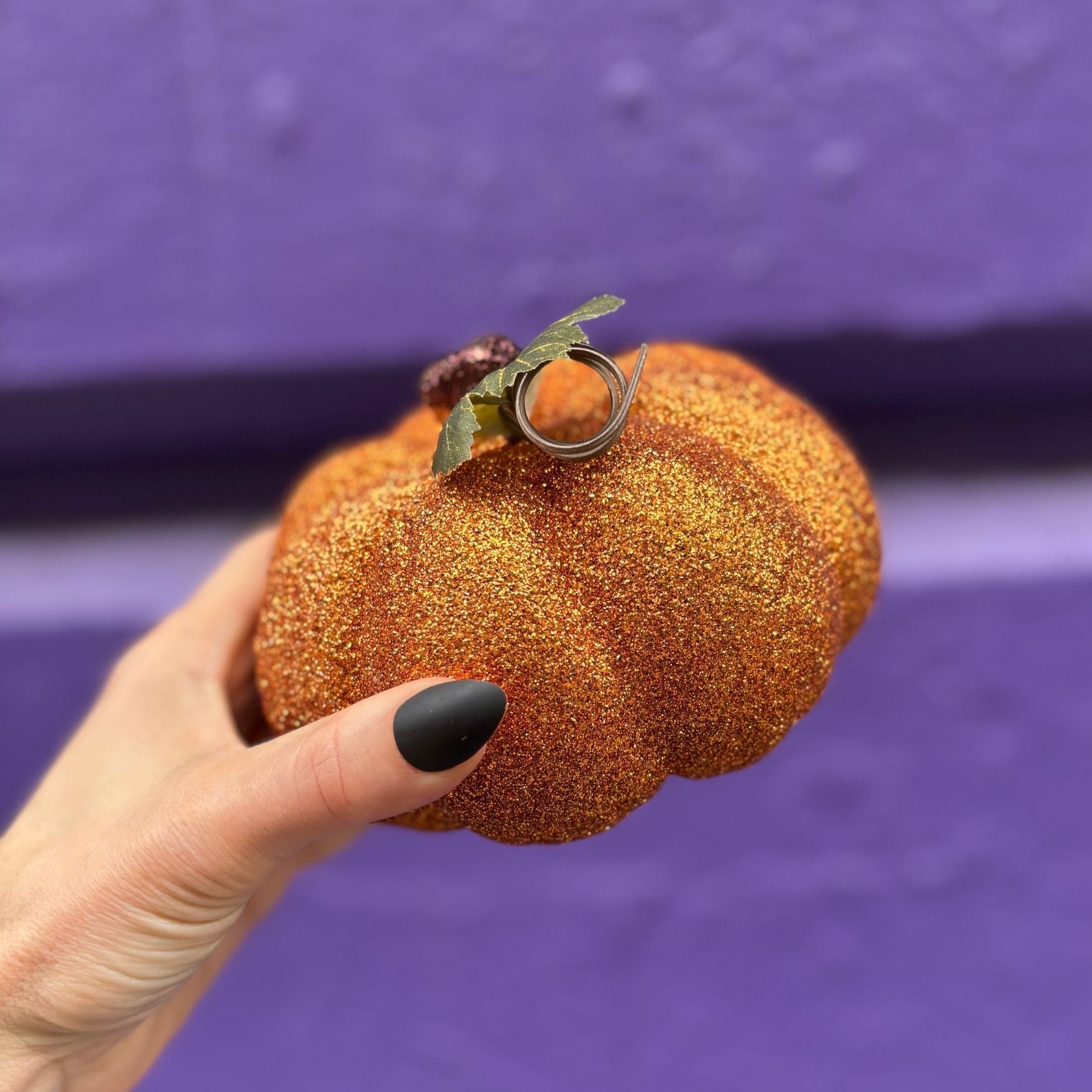 Hand holding a glittery pumpkin-shaped ornament with a ring on a purple background