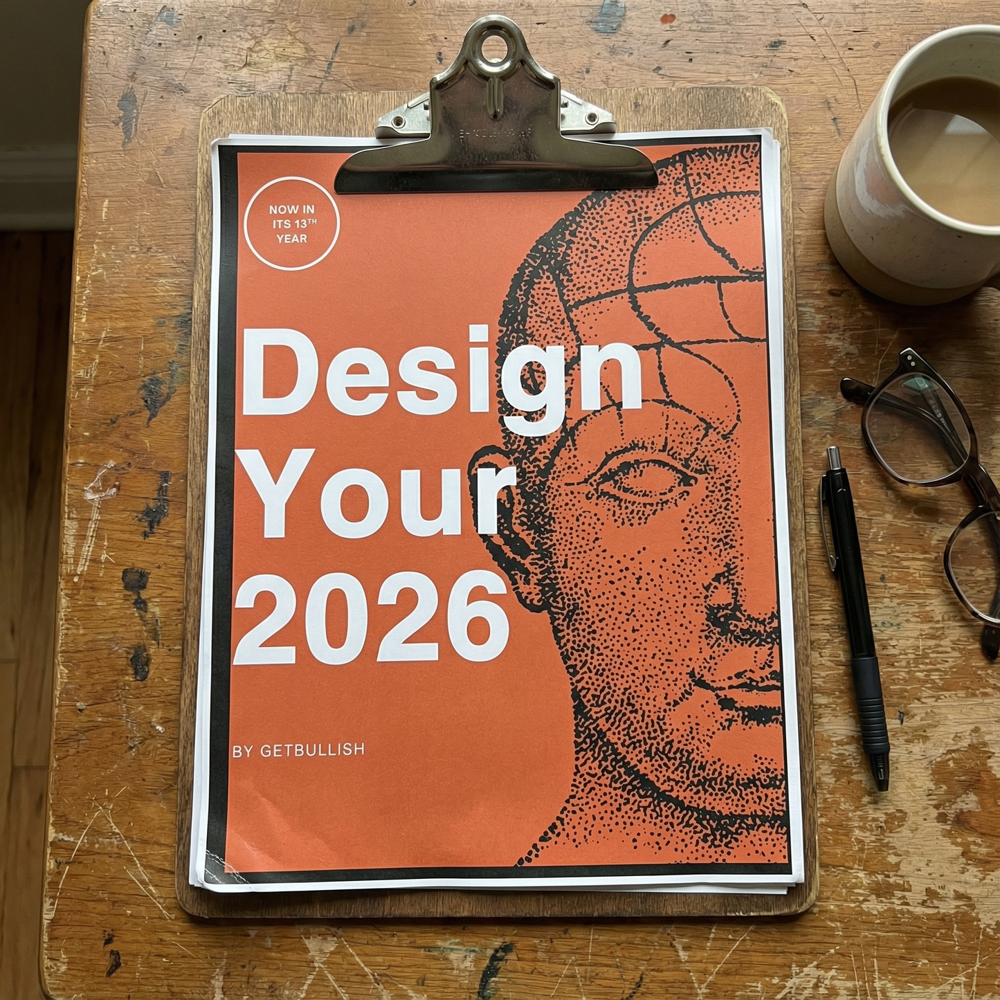 Book titled 'Design Your 2026' on a clipboard with a cup of coffee and glasses on a wooden table.