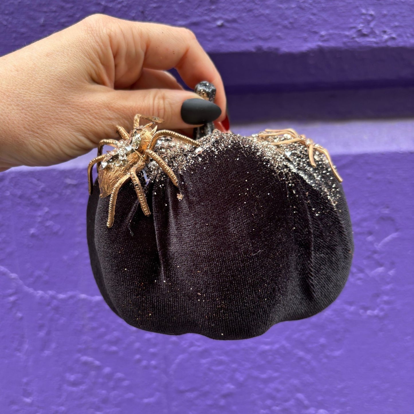 Black glittery pumpkin with spider decorations held by a hand against a purple background
