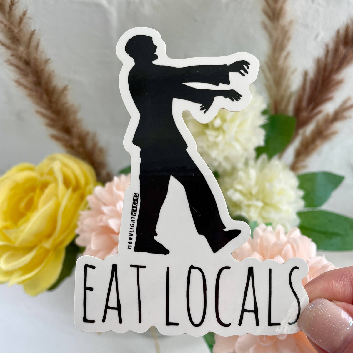 A die-cut vinyl sticker featuring a black silhouette of a person with outstretched arms above the words 'EAT LOCALS' in black text 