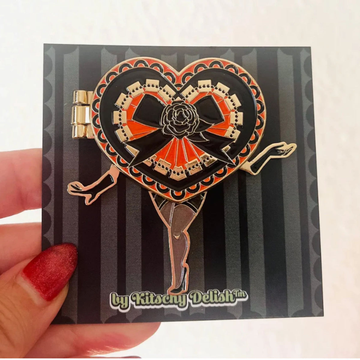 Heart-shaped hinged enamel pin with black and orange design on a striped card, held by a hand with red nail polish.