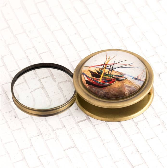 A round brass magnifying glass case with its lid off, showing a detailed painting of colorful fishing boats on the top cover, set against a white brick-patterned background.