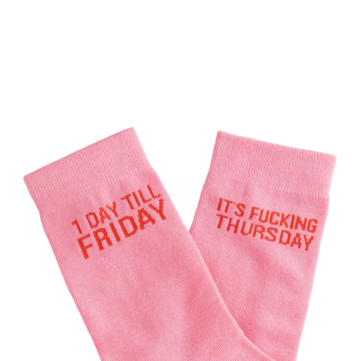 A pair of light pink crew socks with orange-red text. One sock reads '1 DAY TILL FRIDAY' and the other reads 'IT'S FUCKING THURSDAY'