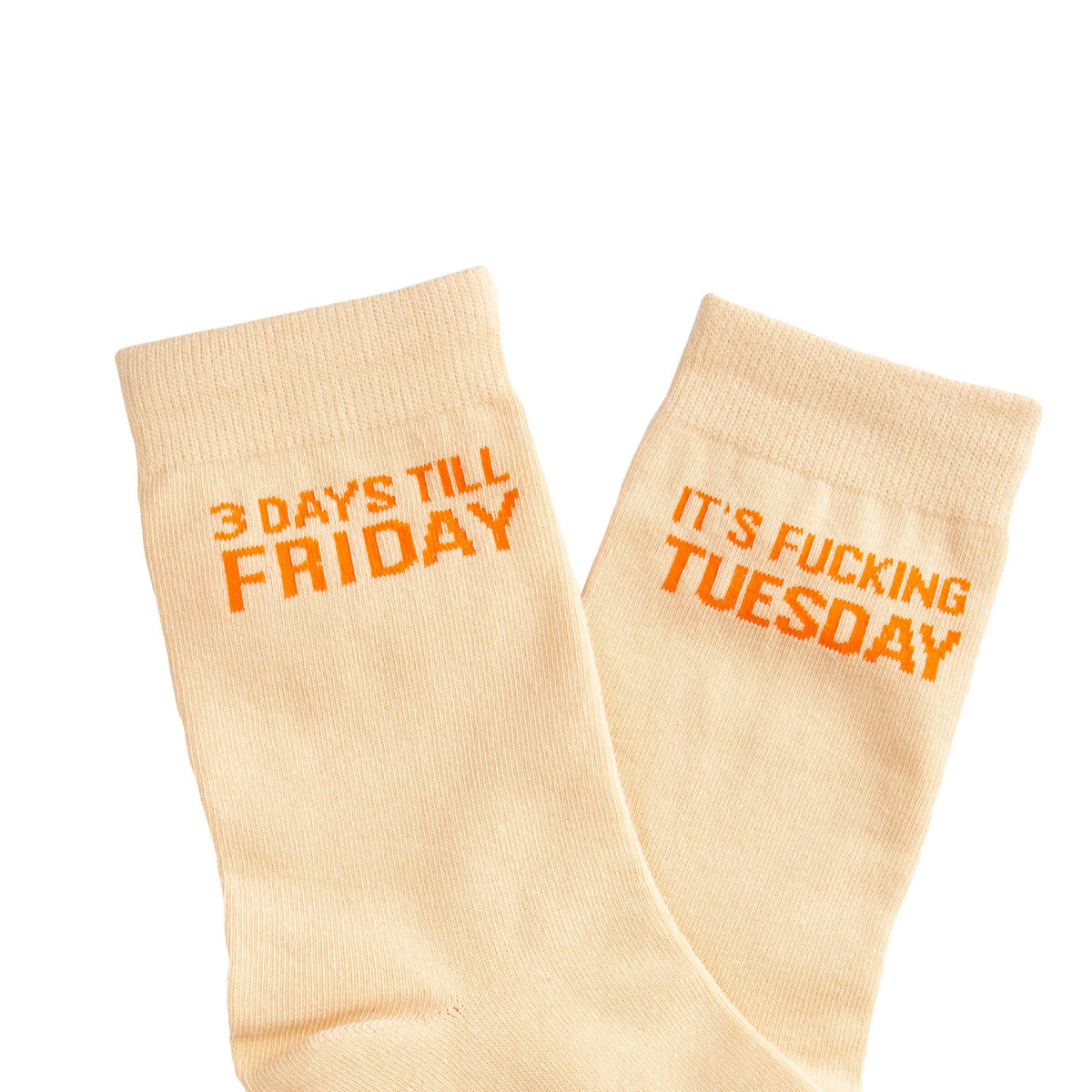 A pair of light beige crew socks with orange text printed on them, one reading '3 DAYS TILL FRIDAY' and the other 'IT'S FUCKING TUESDAY'