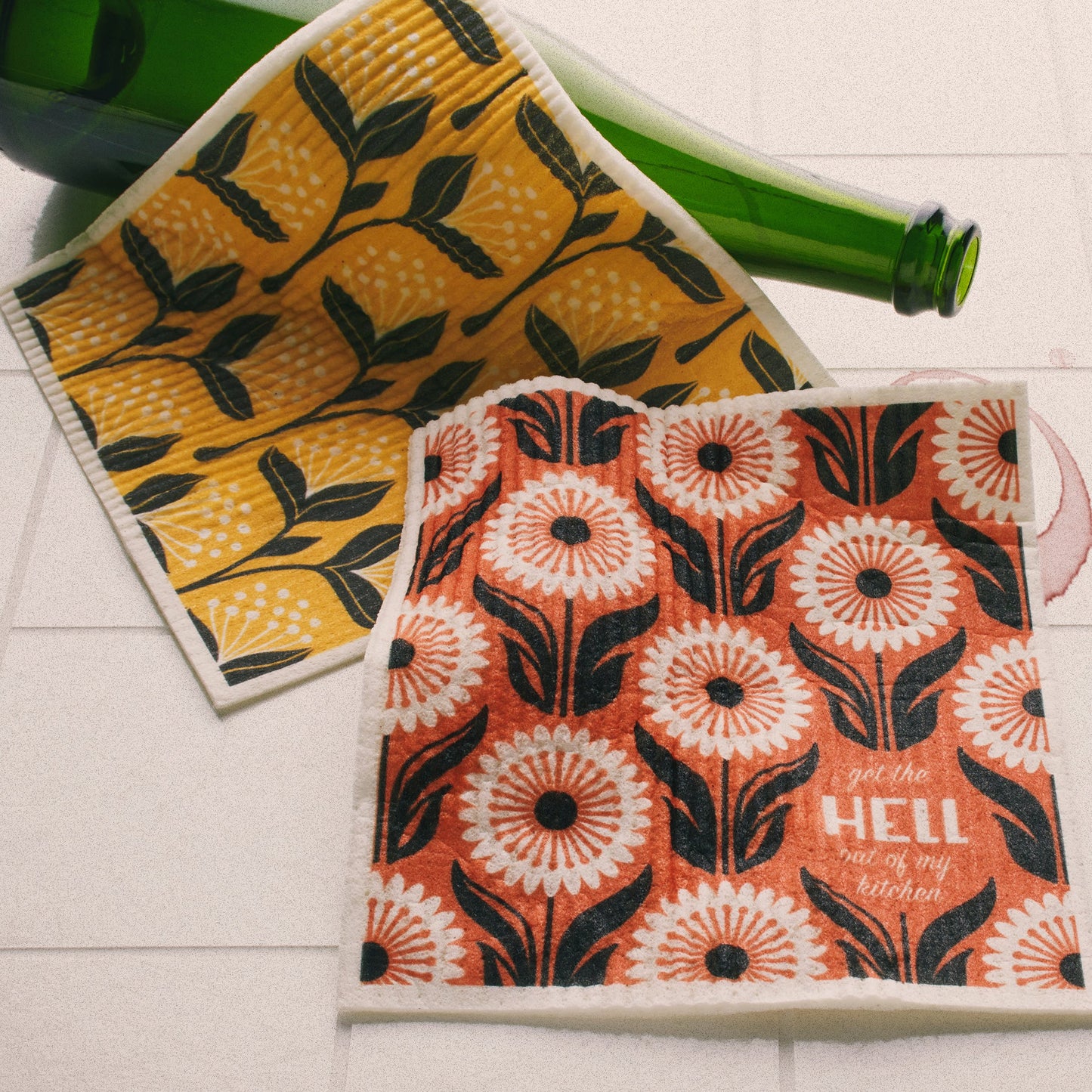 Two patterned dishcloths on a tiled floor with a green bottle.
