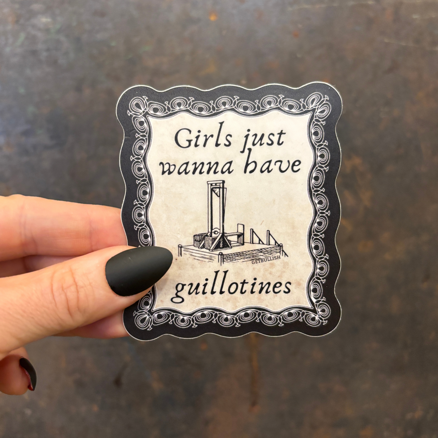 Sticker with text 'Girls just wanna have guillotines' held by a hand on a textured surface