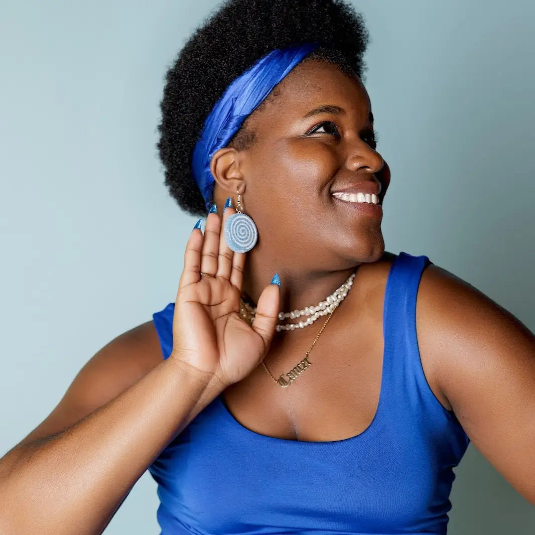 Woman wearing a blue top and headband, holding a blue glitter lollipop earring to her ear