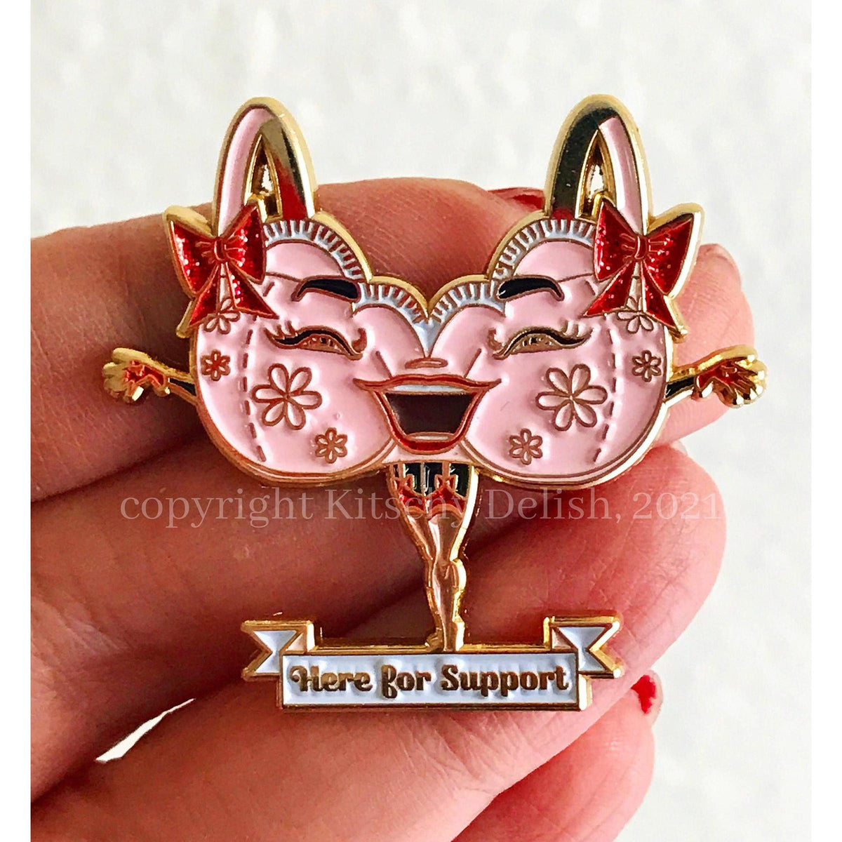 A close-up of a hand holding a decorative enamel pin shaped like two smiling pink faces with gold outlines, red bows, floral details, and a banner reading 'Here for Support' at the bottom.