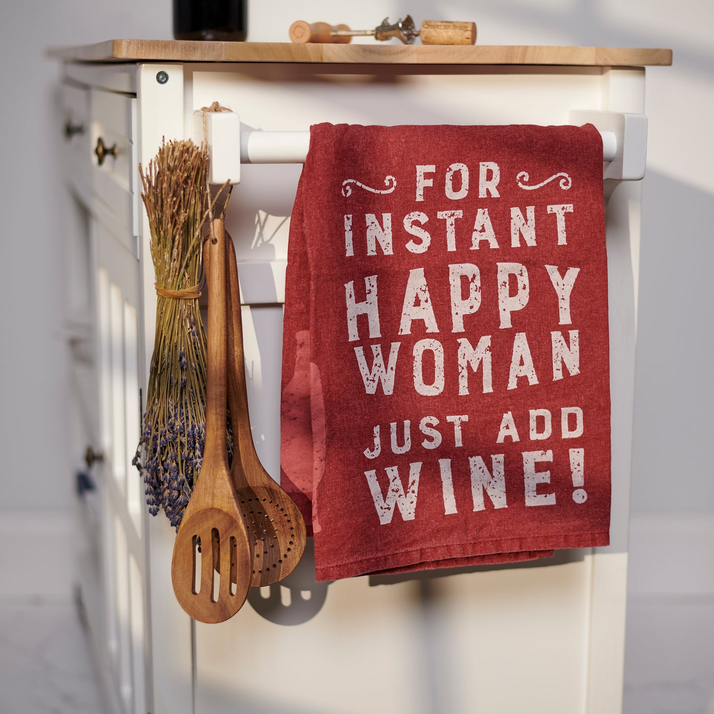 Red towel with humorous text hanging on a wooden cabinet with kitchen utensils.