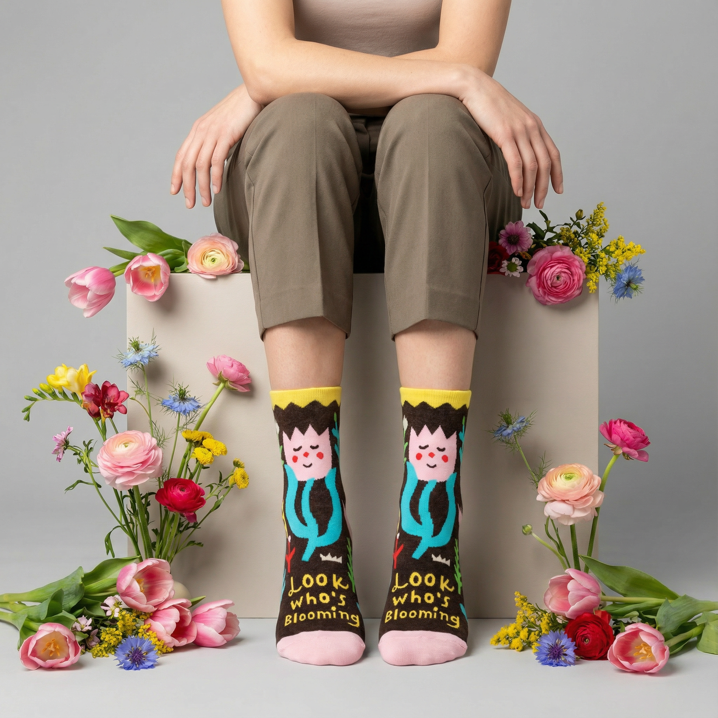 Person wearing socks with cartoon characters and text, sitting on a box with flowers around.